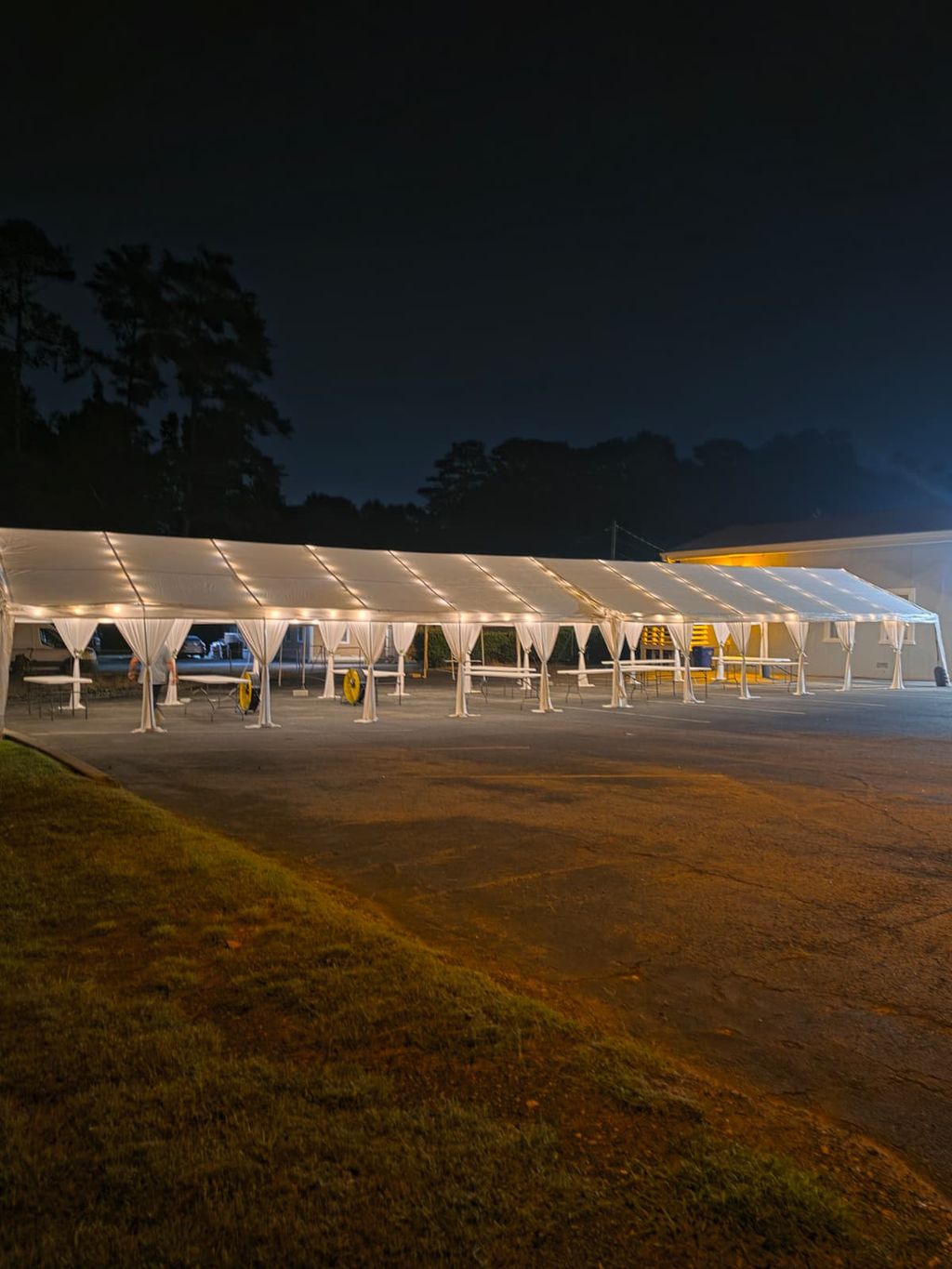 Long white frame tent with draped legs and warm string lights lit at night on a paved lot — event setup in Avalon District, Alpharetta, GA