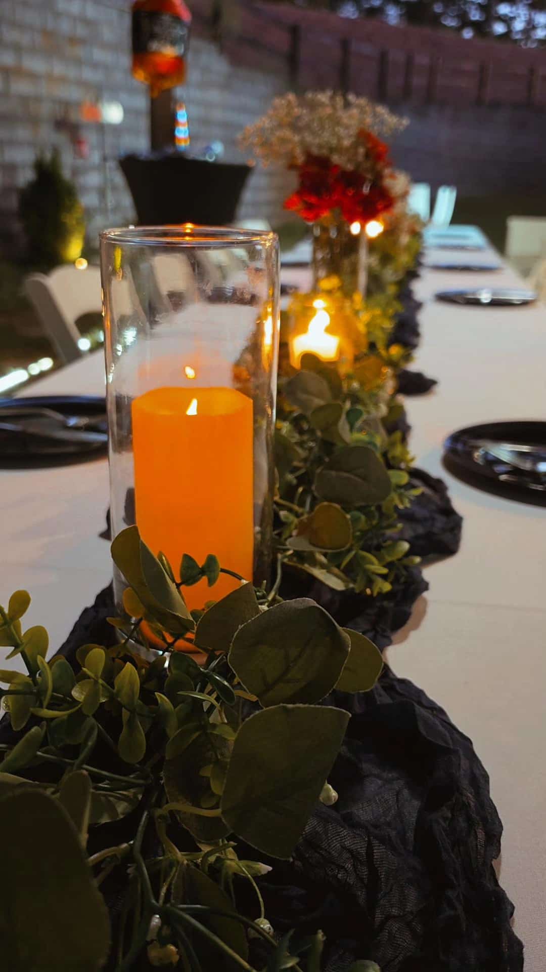 Pillar candle in a glass hurricane with eucalyptus and red rose centerpieces along an evening banquet table