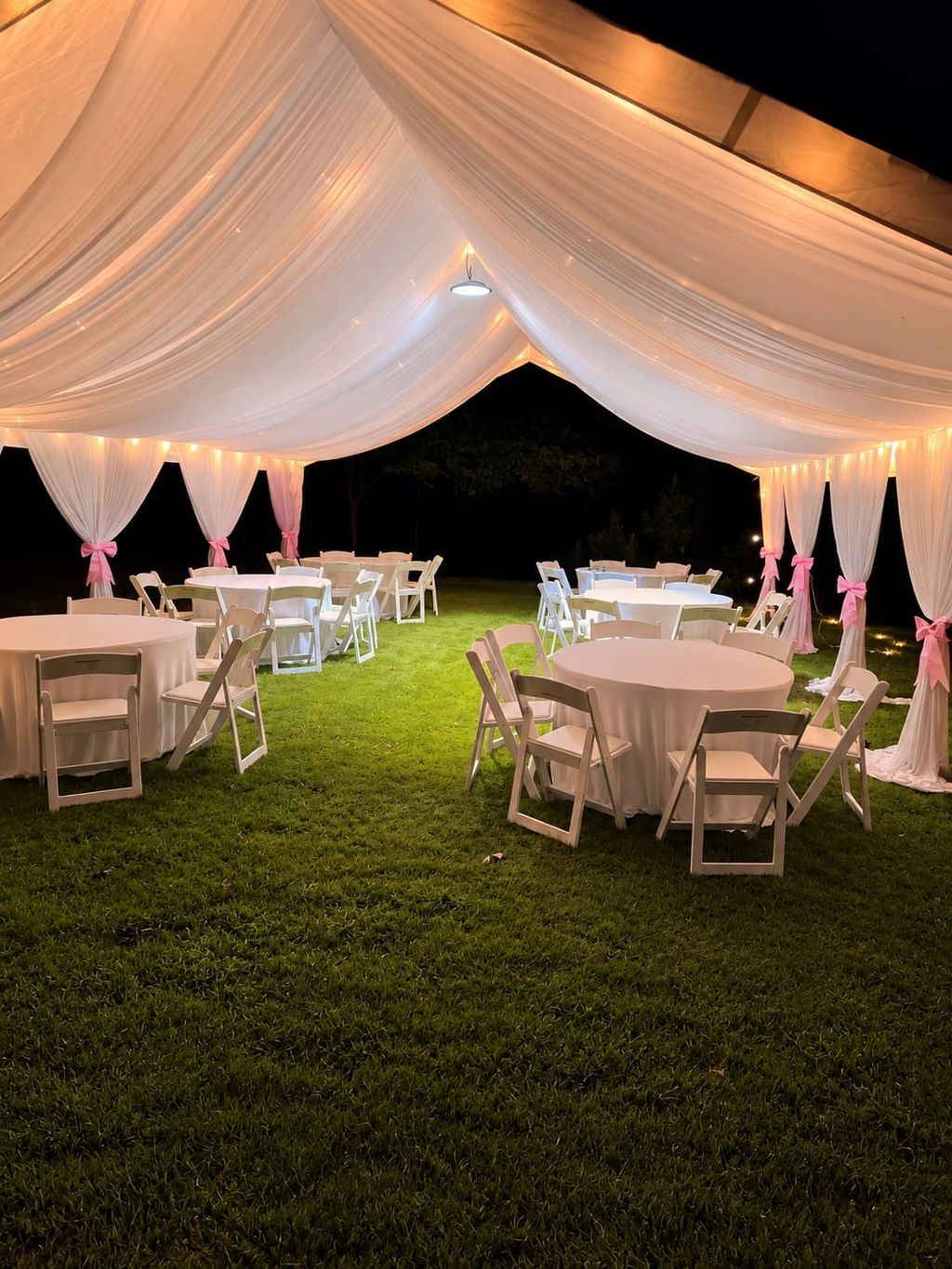 Draped white tent with pink bows and a round table with white folding chairs on grass at night