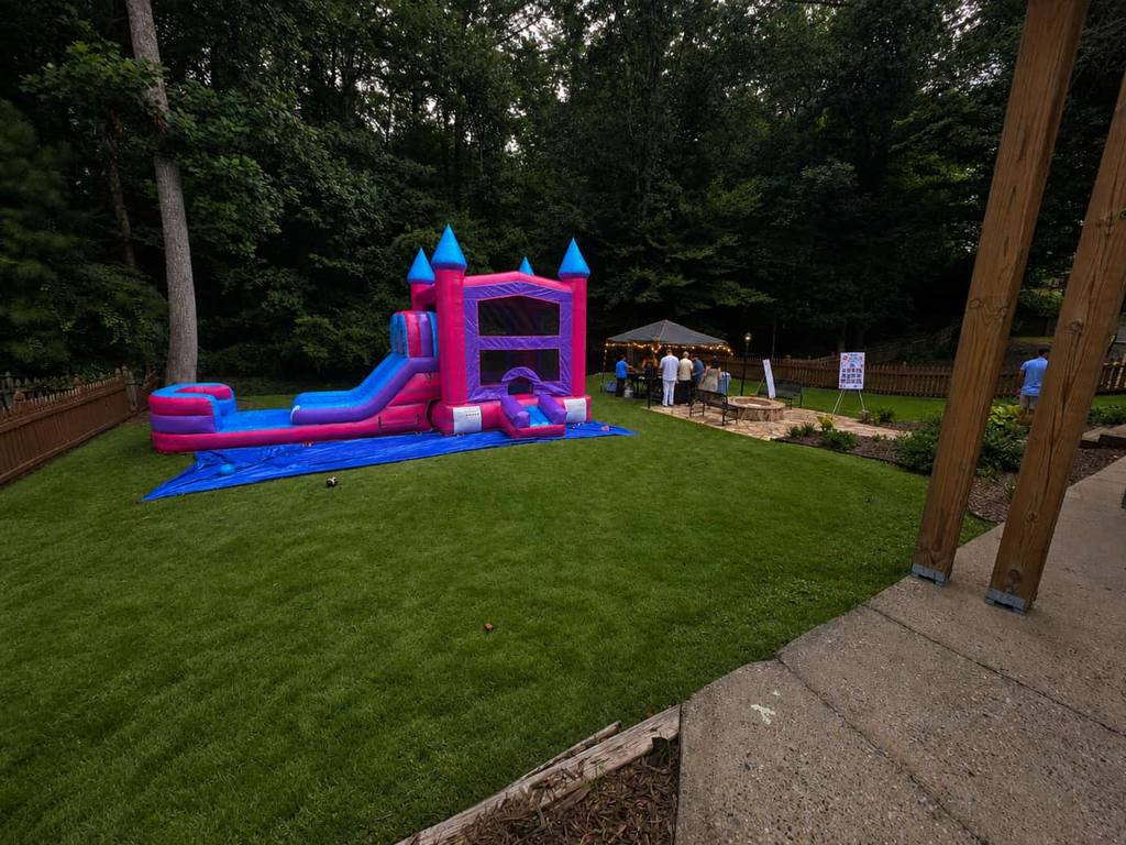 Pink and blue castle bounce house with water slide on a manicured backyard lawn beside a lit event tent