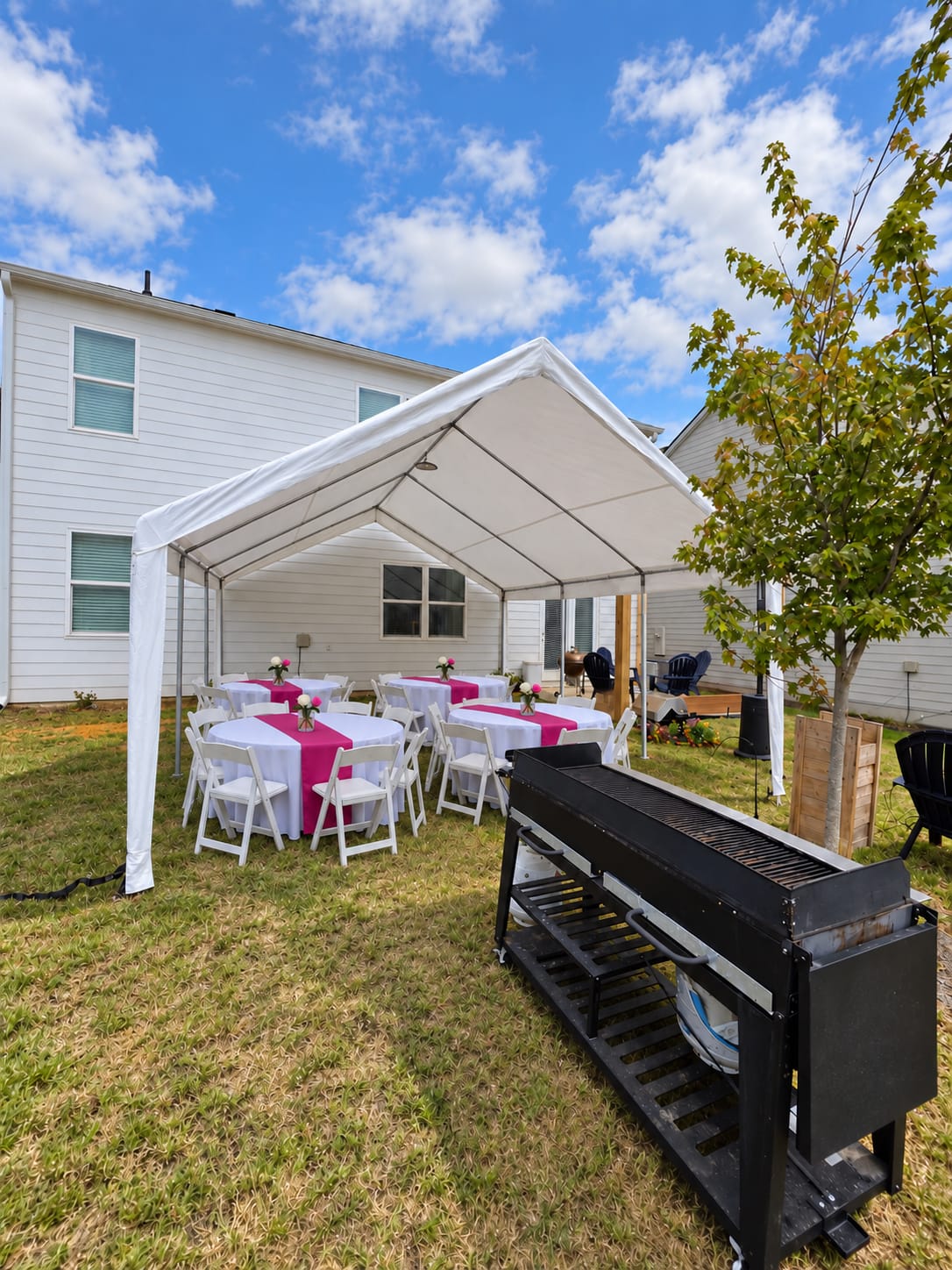 Backyard frame tent with round tables, pink runners and white folding chairs beside a grill station