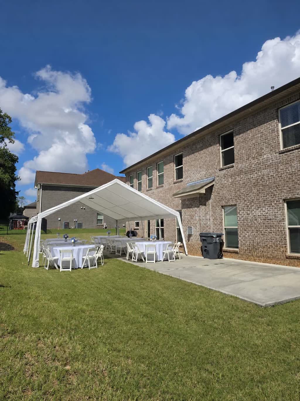 Frame tent with white tables and chairs beside a brick home on a sunny afternoon