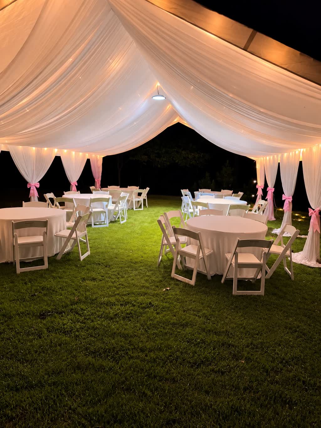 Draped white tent glowing at night with round tables, white folding chairs and pink sashes