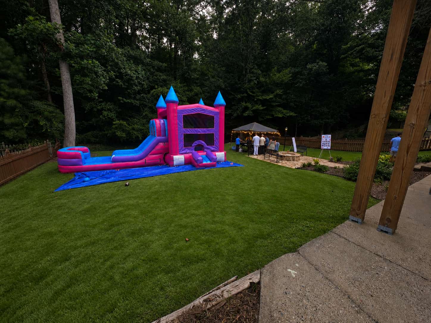Pink and blue castle bounce house with water slide on a manicured backyard lawn beside a lit event tent