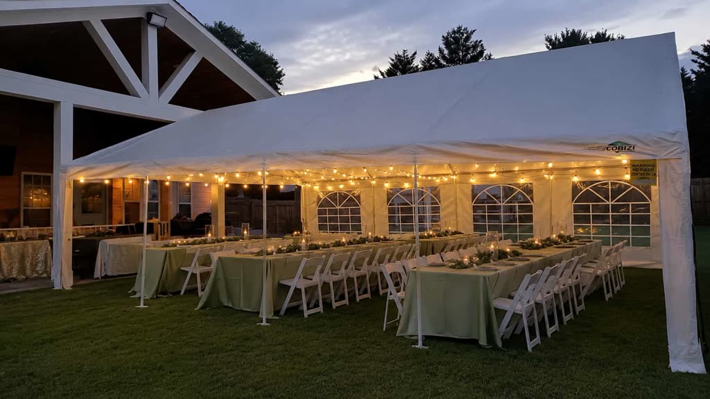 Long white tent with sidewall windows, string lights and sage green banquet tables set for dinner at dusk