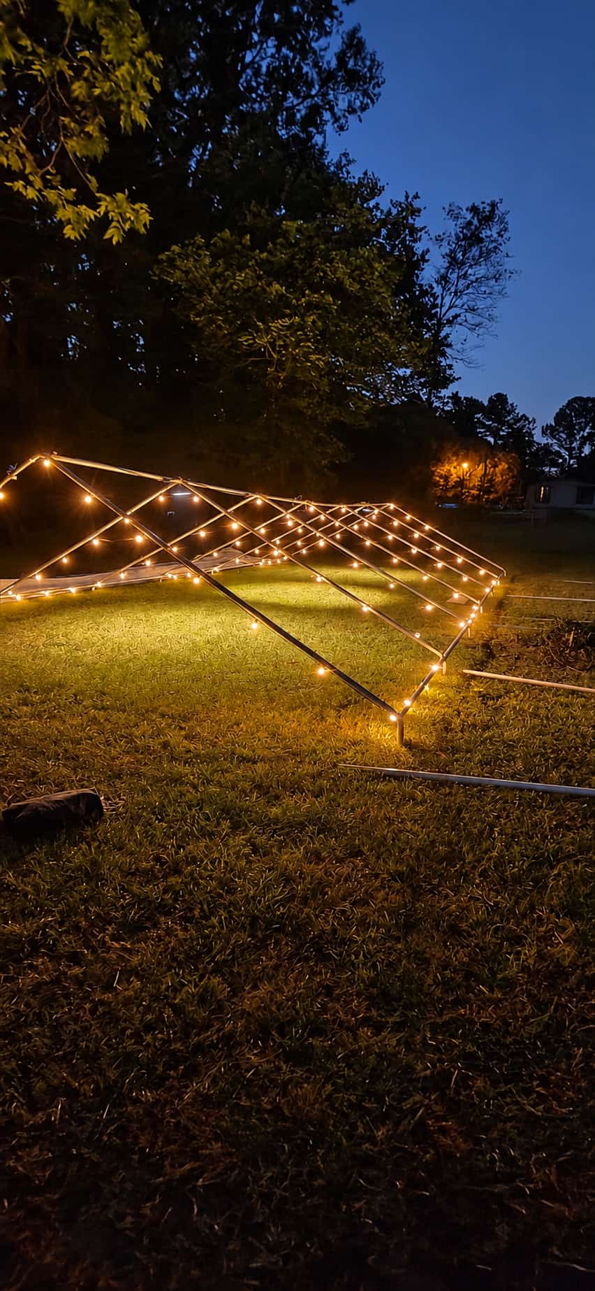 Tent frame wrapped in warm string lights glowing on a lawn at night before the canopy goes on