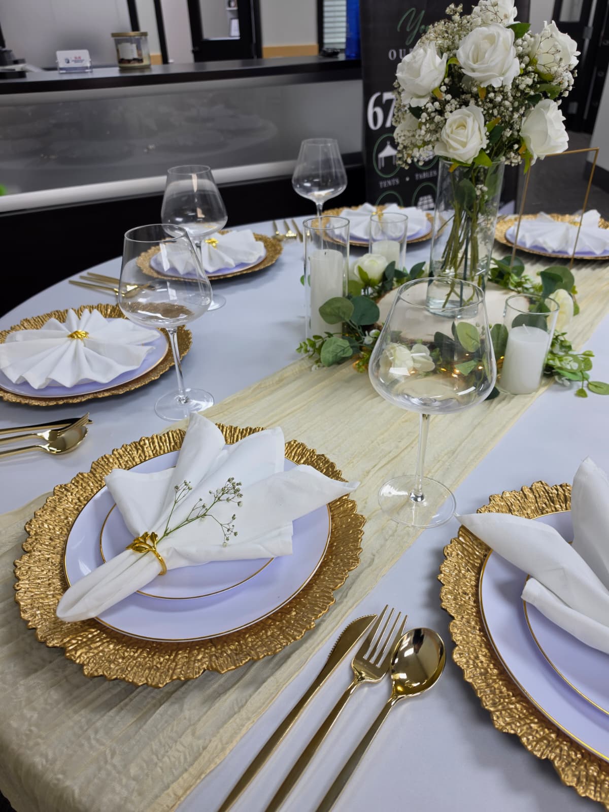 Round table set with gold chargers, gold flatware, knotted white napkins, wine glasses and a white rose centerpiece