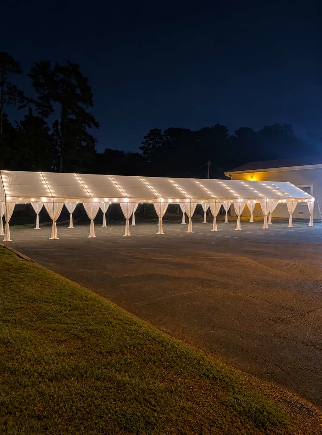 Draped white tent with pink bows, round tables and white folding chairs on a lawn at night — event setup in Avalon District, Alpharetta, GA