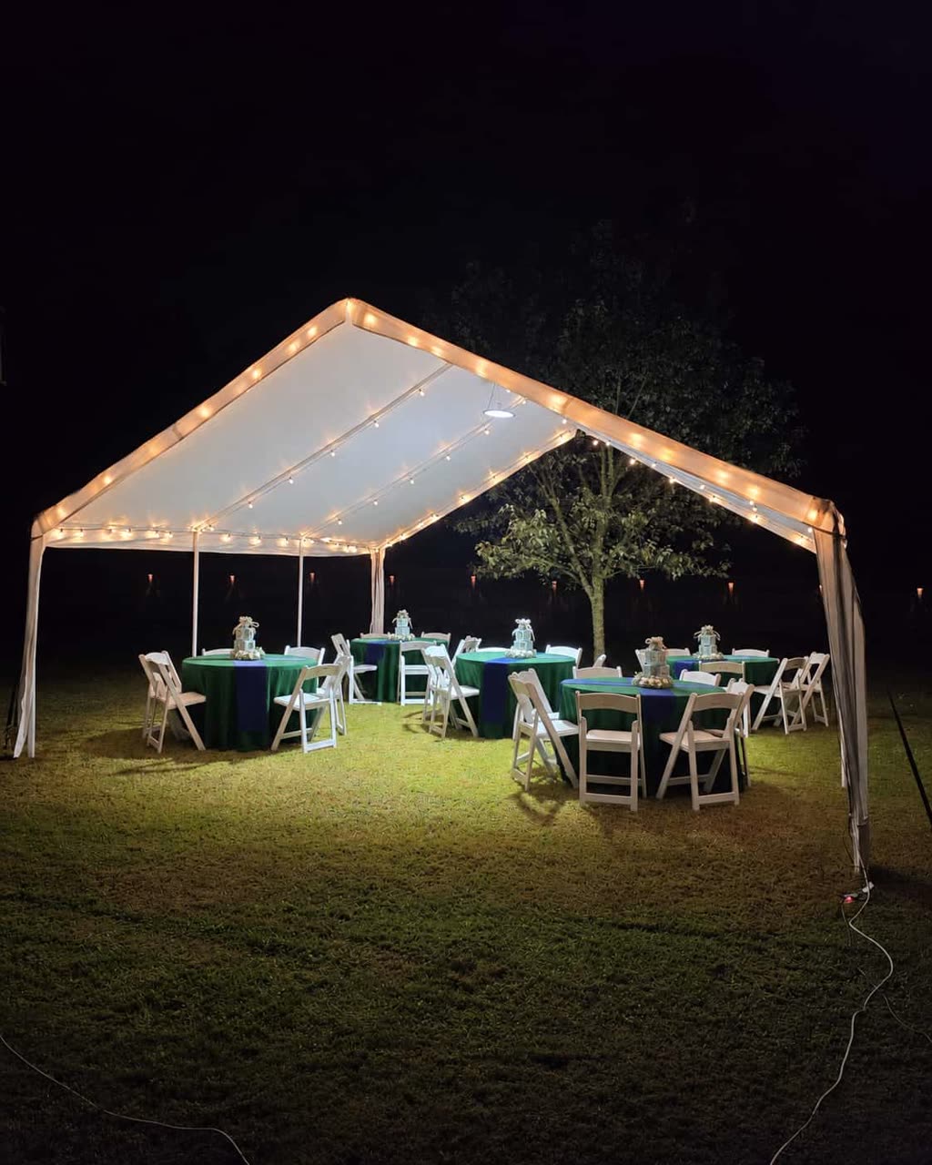 White frame tent with round tables, ivory runners and folding chairs under shade trees